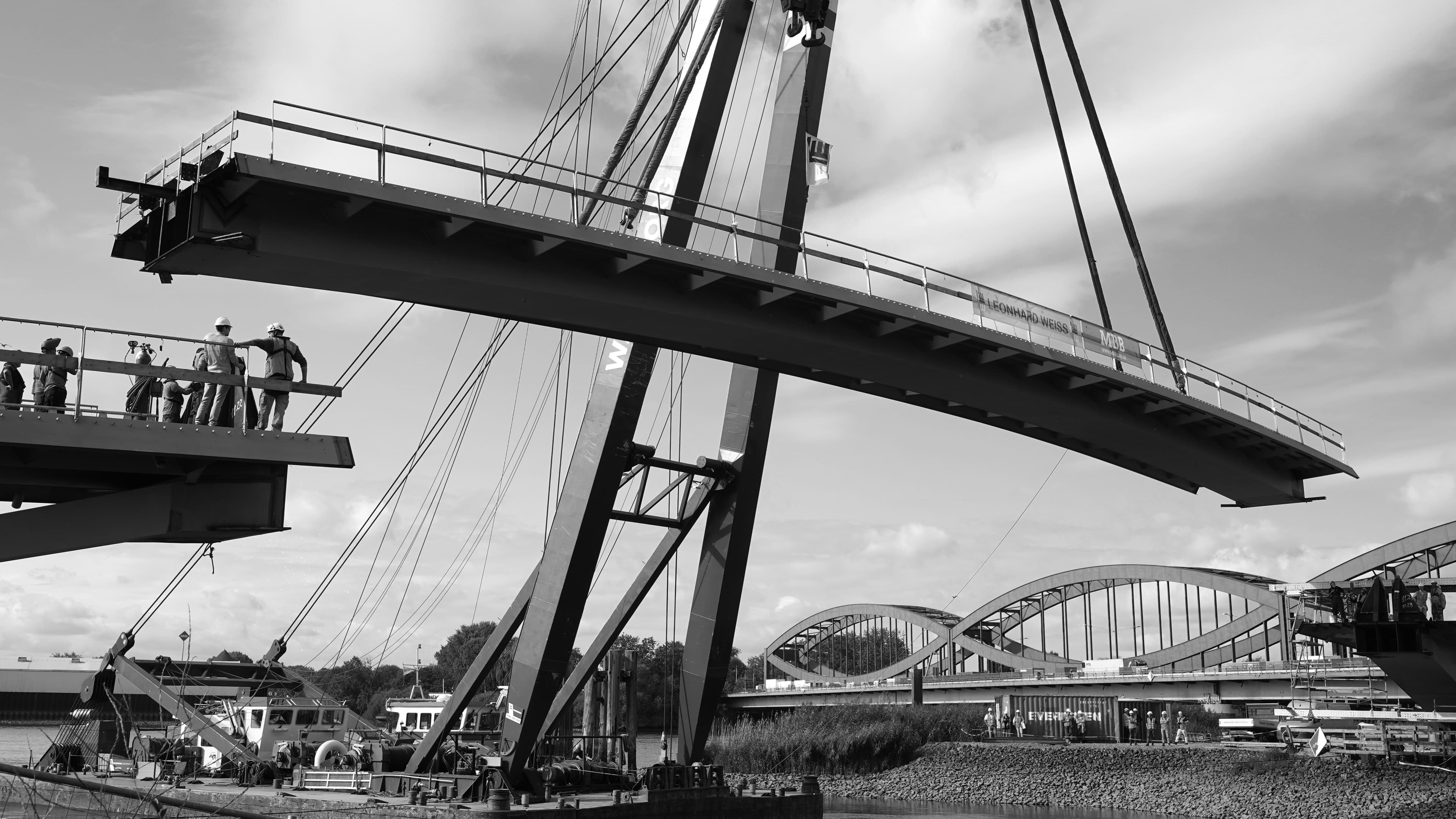A steel bridge span being lifted into place by crane, connecting two banks